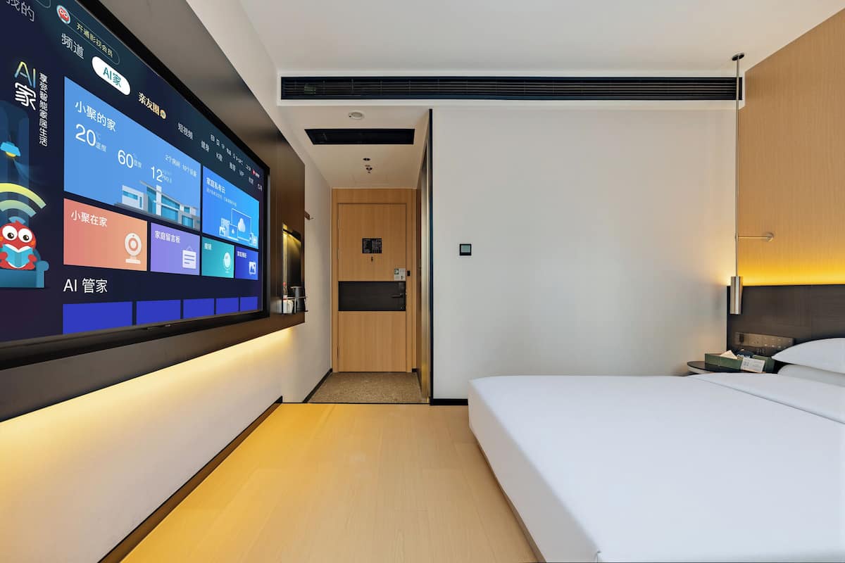 A modern hotel room with a large digital display on the wall, a bed, bedside table, and a wooden door leading to the bathroom.