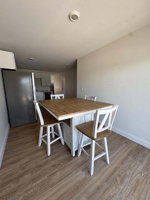 Un coin repas avec une table en bois et des chaises blanches, une cuisine équipée d’un réfrigérateur et un mur de couleur claire.