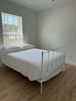 Une chambre avec un lit simple, un cadre en métal, du linge de lit blanc et une fenêtre avec des stores.