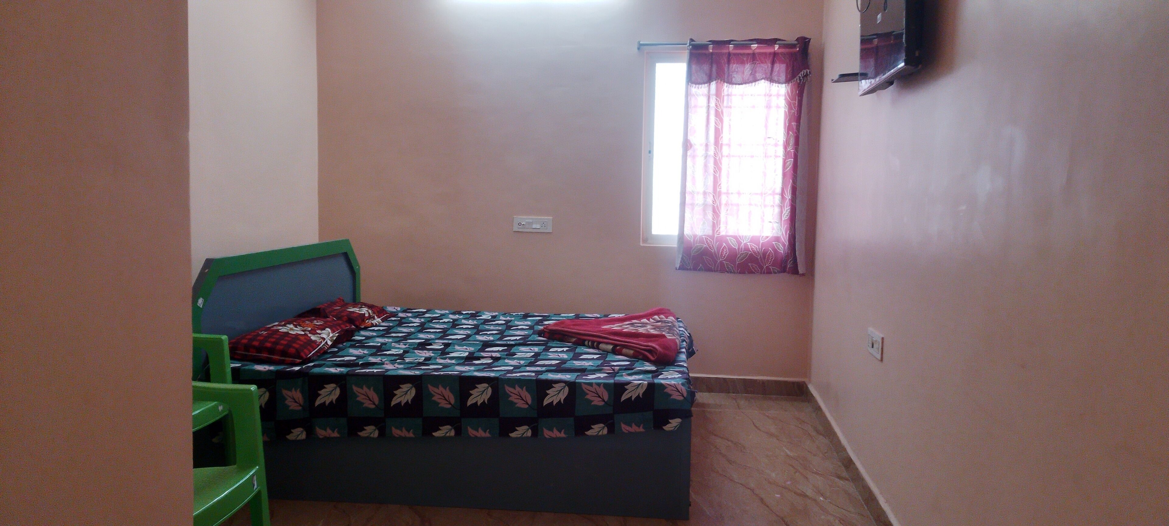 A small bedroom with a bed, a window with curtains, a green chair, and a wall-mounted TV.