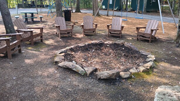 A campsite with a central fire pit surrounded by Adirondack chairs, picnic tables, and a playground in the background.