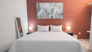 Un letto rifatto con lenzuola bianche, due abat-jour, un grande quadro astratto sulla parete e uno specchio a sinistra.
