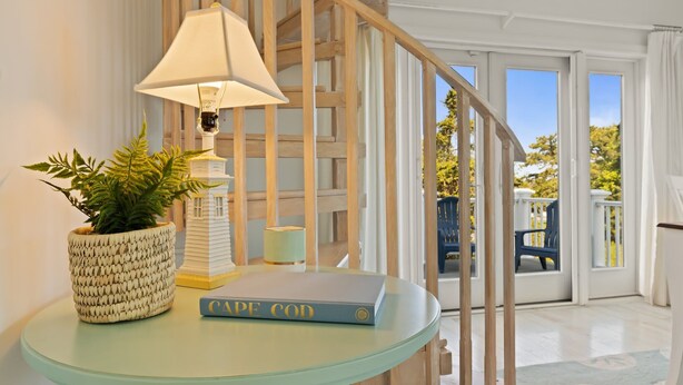 Um quarto com uma mesa curva, uma luminária, um livro, uma planta em vaso e vista para o exterior através de uma porta de vidro.