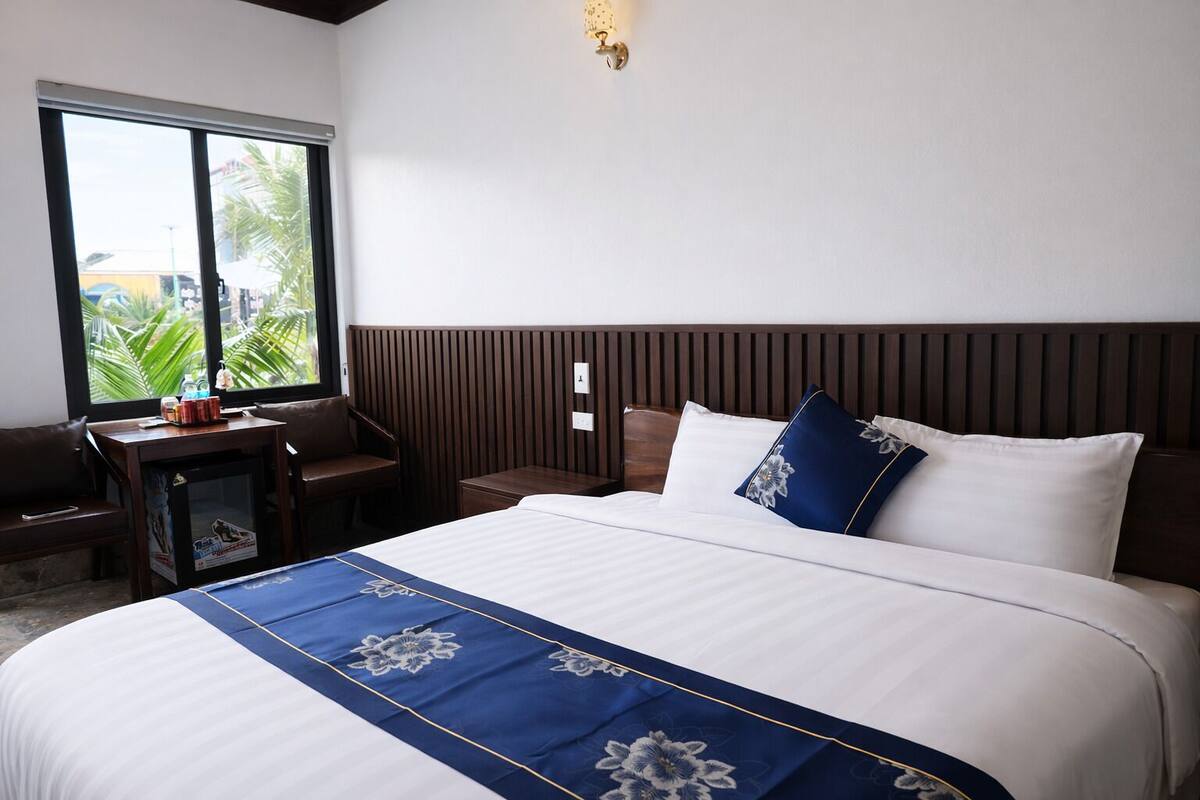 A hotel room with a large bed, a wooden headboard, a small table with a chair, and a window with a view of palm trees.