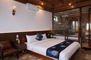 A hotel room with a large bed, a wooden headboard, a nightstand, a brown leather chair, and a glass shower enclosure. - Senna Garden HomeStay (Hoa Lu)