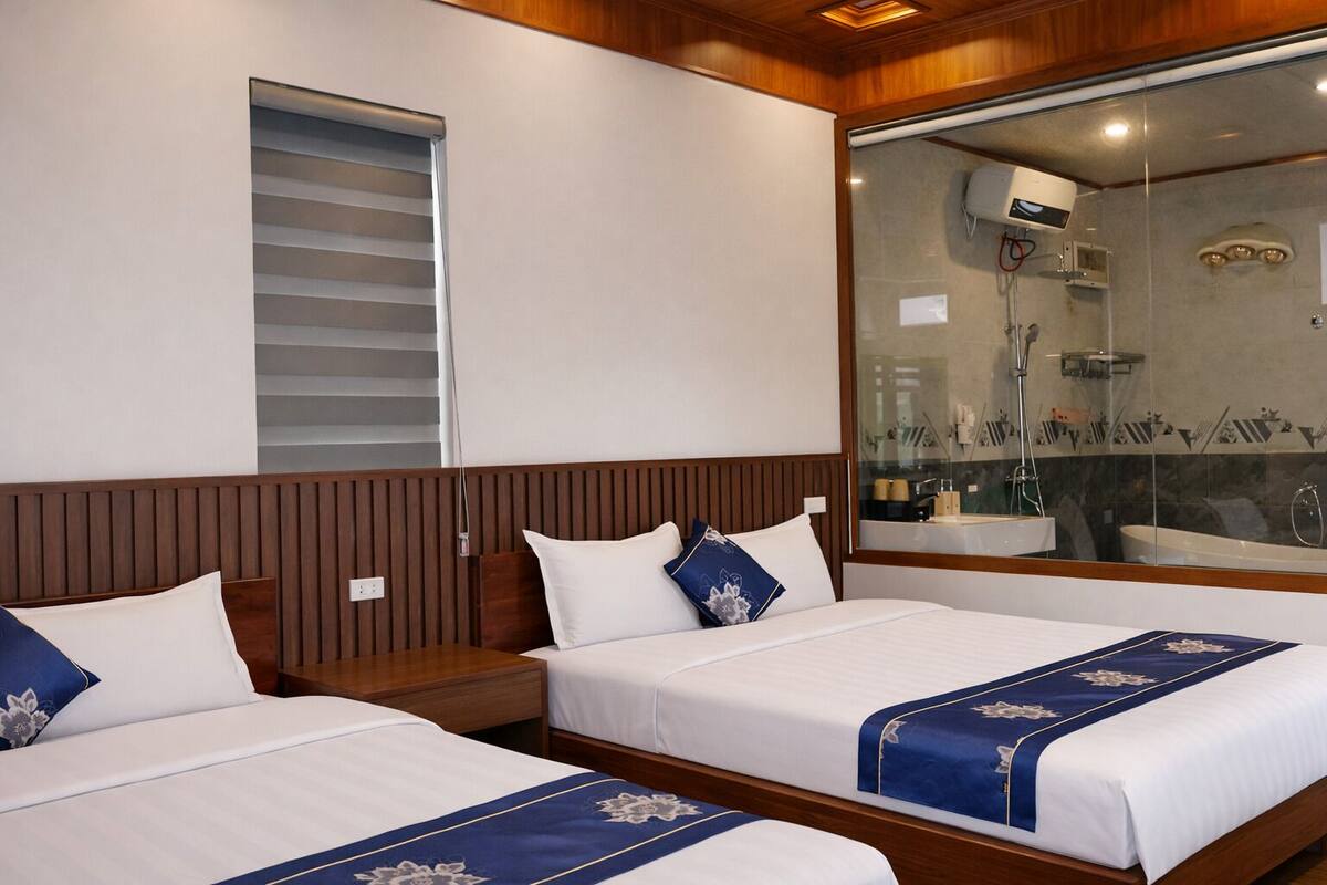 A hotel room with two beds, a large window with blinds, a shower area with a glass partition, and wooden paneling on the walls.