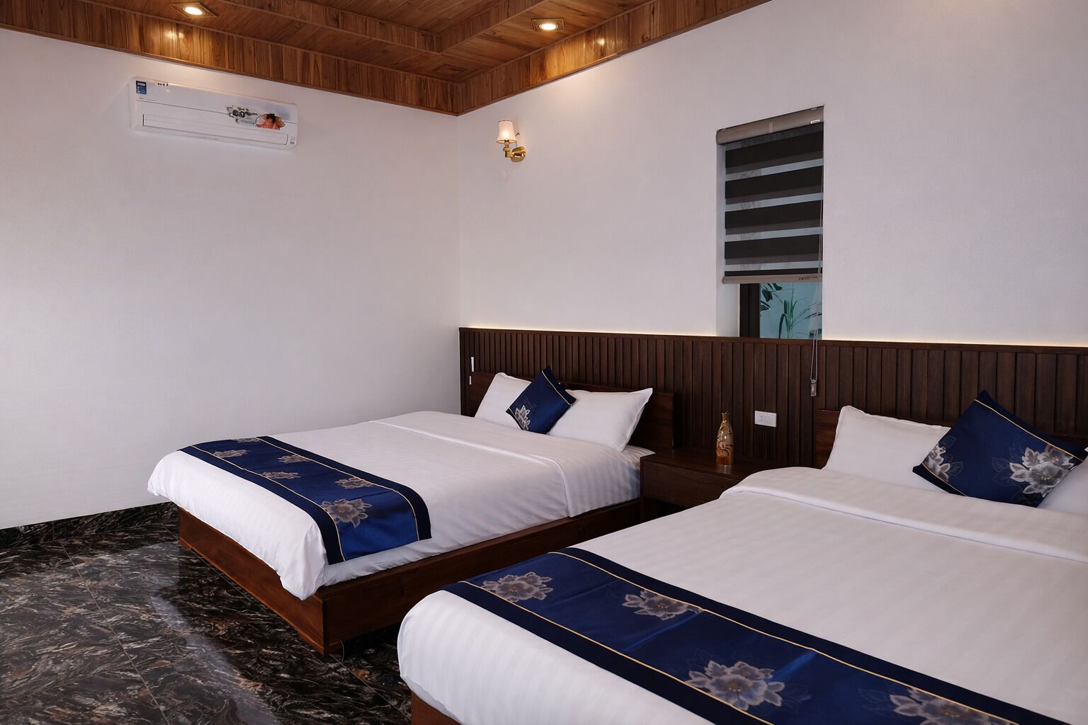 A hotel room with two beds, wooden headboards, a wall-mounted air conditioner, a wall-mounted lamp, and a window with blinds.