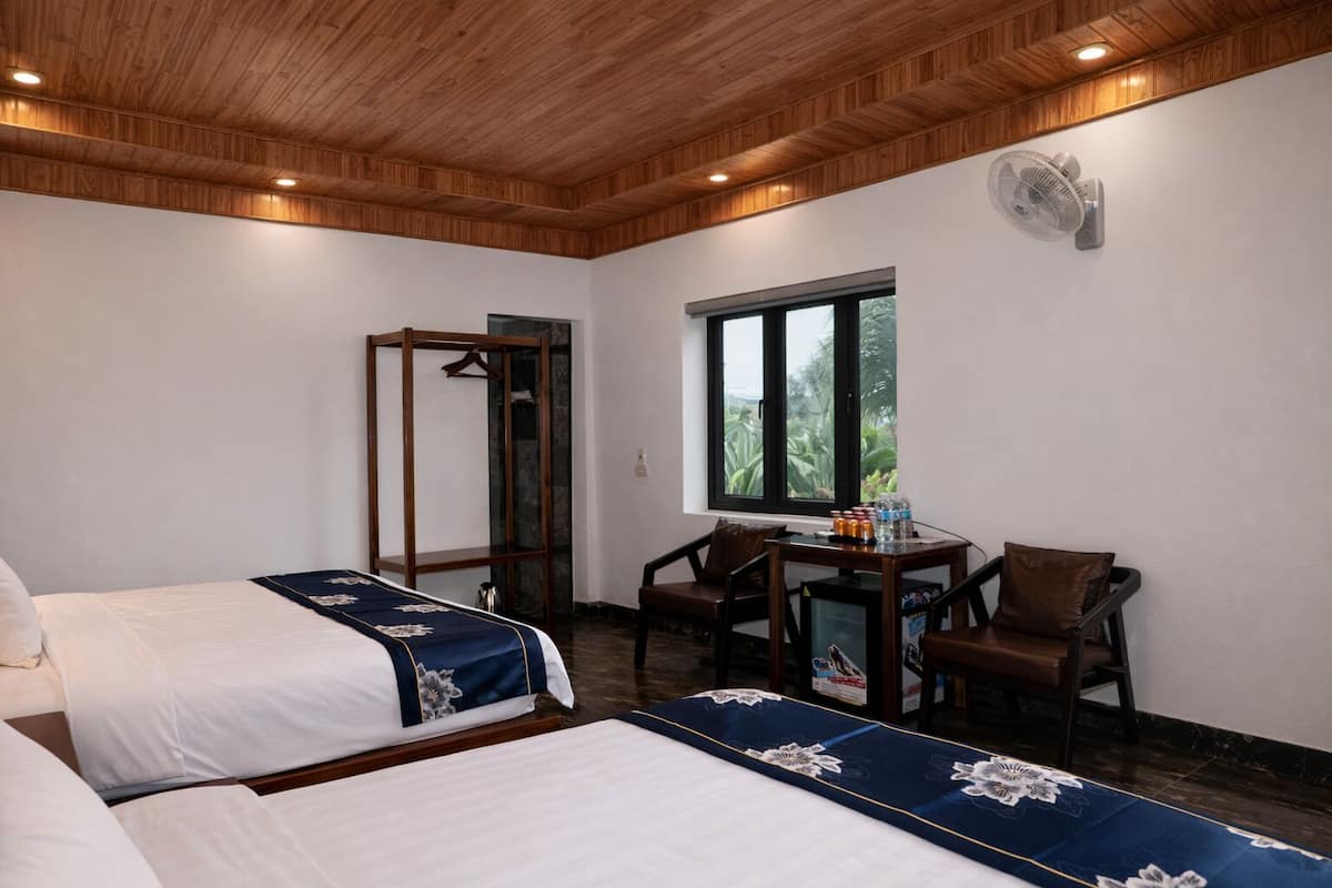 A hotel room with two beds, a wooden ceiling, a window with a view, a table with chairs, and a fan.