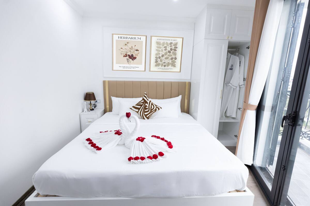 Un lit bien fait, agrémenté d’une décoration représentant un cygne et une rose, une table de chevet avec une lampe, et une armoire où sont suspendus des vêtements.