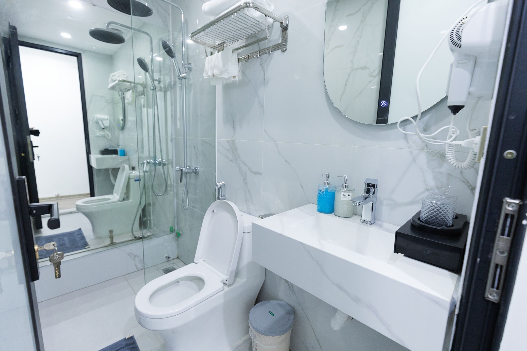 A modern bathroom with a glass-enclosed shower, a white toilet, a sink with toiletries, and a hairdryer on the wall.