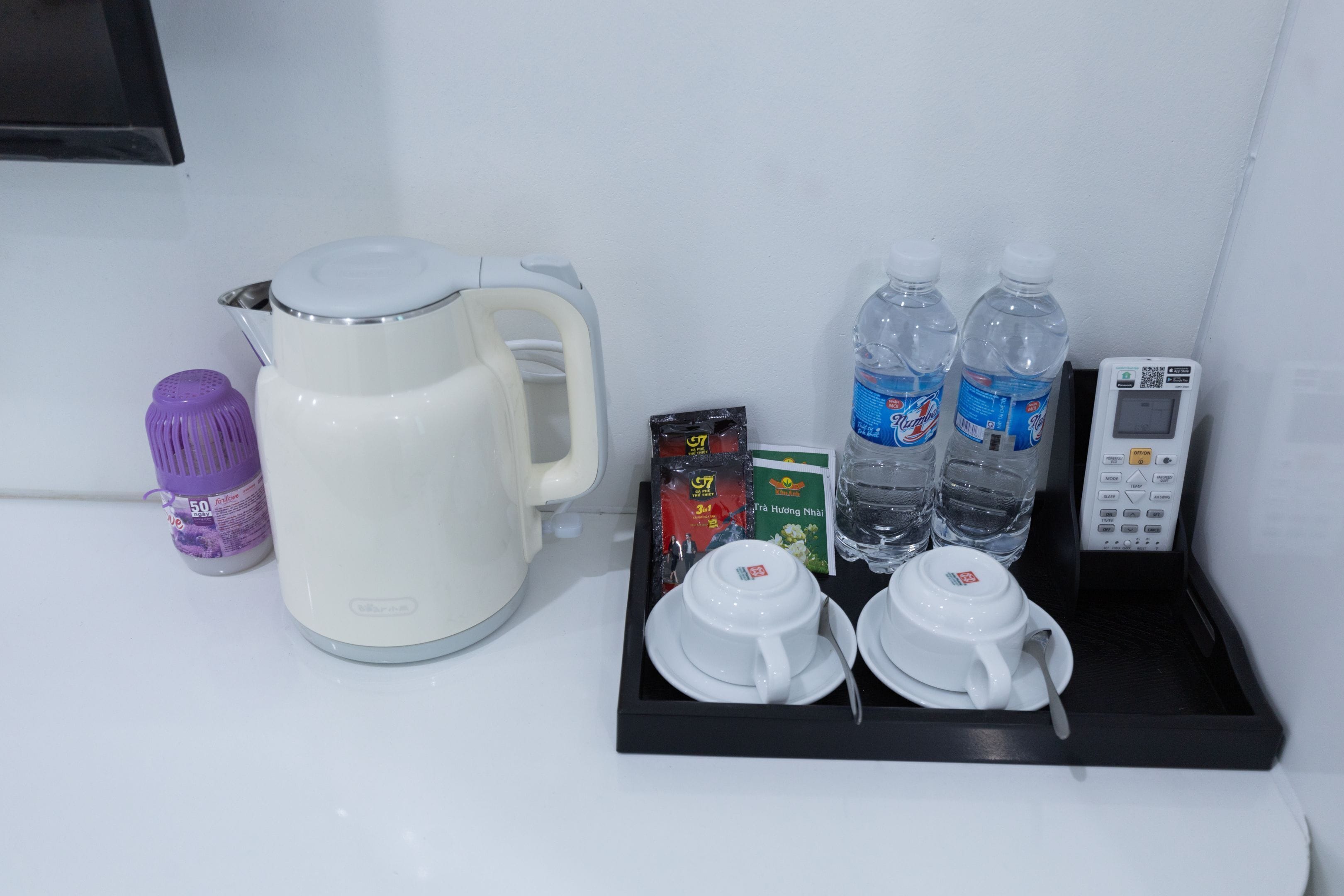 Eine Kaffeemaschine, Wasserflaschen, ein Wasserkocher, ein Tablett mit Tassen und einem Löffel, eine kleine violette Flasche und ein QR-Code-Scanner auf einer weißen Oberfläche.