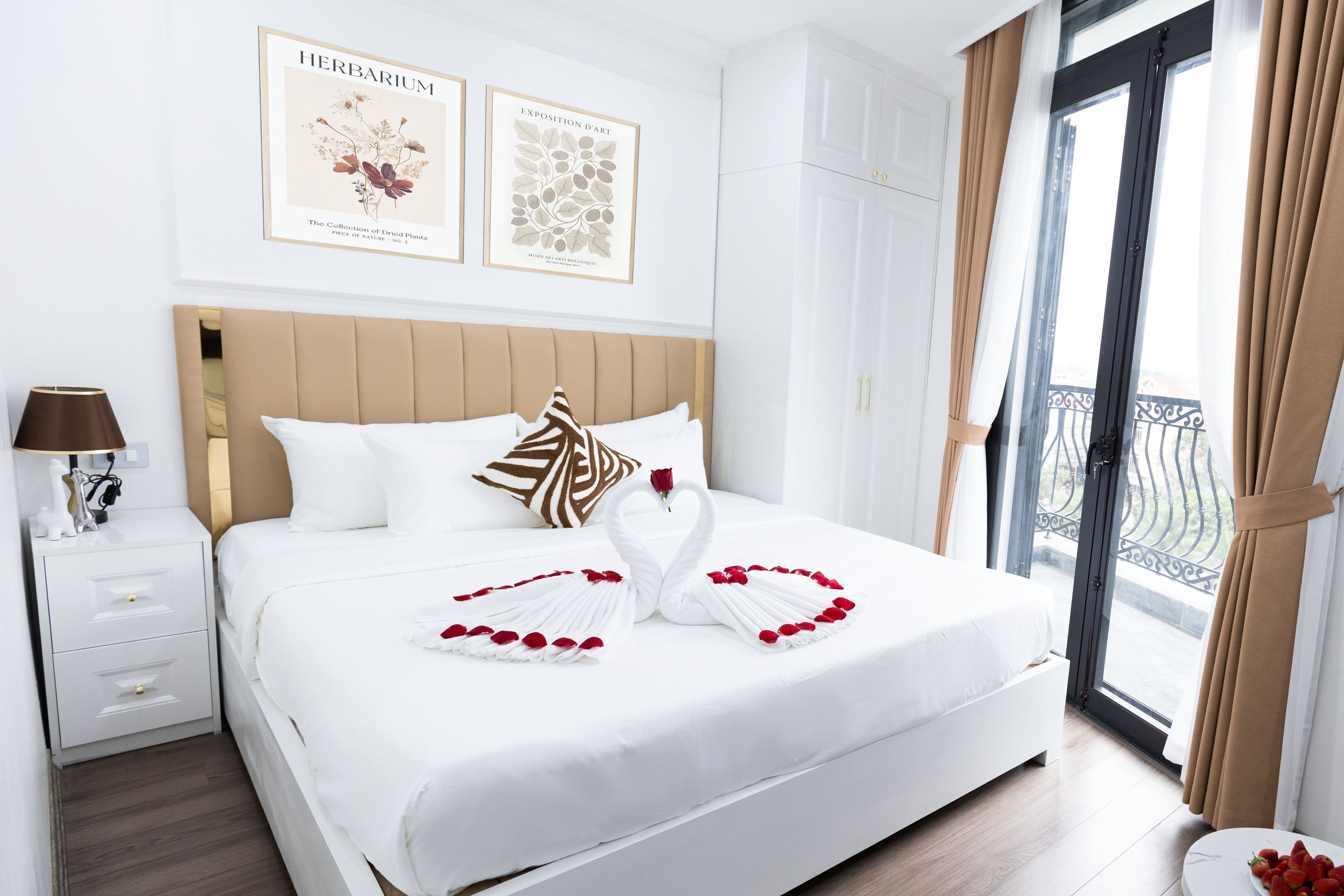 A hotel room with a large bed, a nightstand with a lamp, a framed botanical print on the wall, and a balcony with a view.