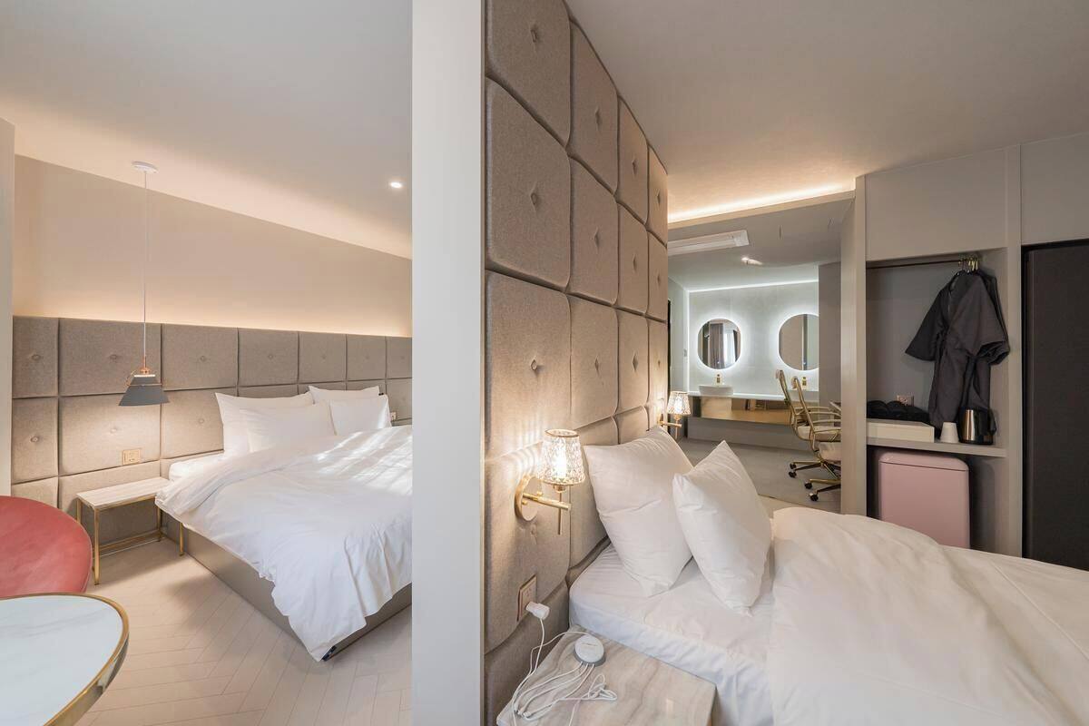 A modern hotel room with two beds, a red chair, a desk, and a vanity with mirrors.