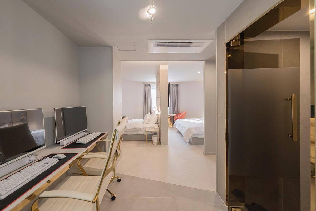 A hotel room with a desk, two monitors, a chair, a bed, and a bathroom.
