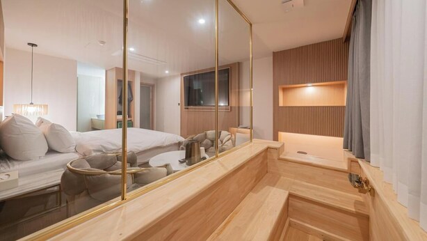A modern hotel room with a large mirror, a wooden bench, a bed with pillows, a bedside table, and a TV. - Denbasta Hotel Busan Jeonggwan (Busan)