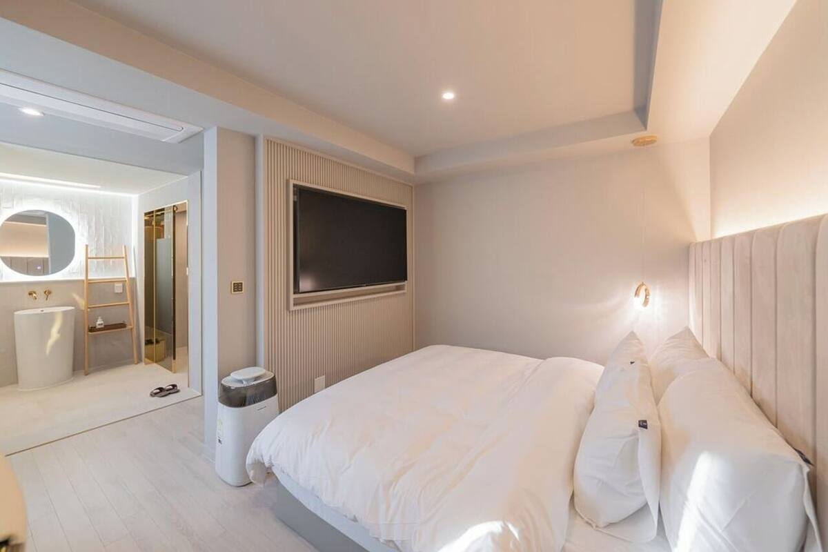 A modern bedroom with a large bed, a TV mounted on the wall, and a bathroom visible through an open door.