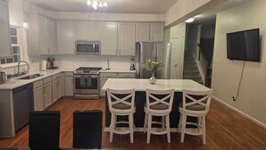 A modern kitchen with white cabinets, stainless steel appliances, a dining area with a white table and chairs, and a flat-screen TV mounted on the wall. - 5-BR / King Beds / Massive Loft / Amazing Location / 10 mins to Downtown Raleigh (Knightdale)