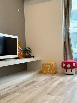 Ein Zimmer mit Fernseher auf einem weißen Regal, einer kleinen Pflanze, einem Mario-Spielzeugauto, einem gelben Block mit einem Fragezeichen und einem pilzförmigen Kissen.