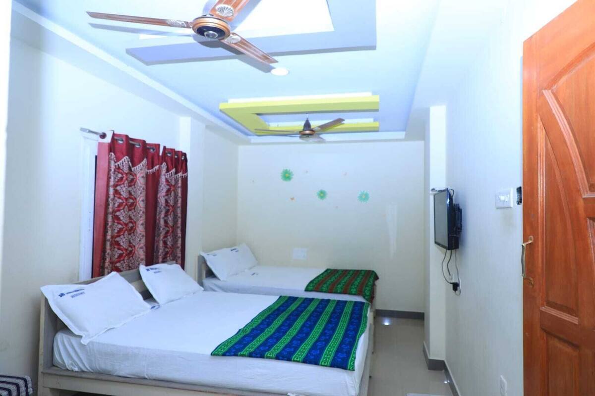 A small, well-lit room with two single beds, a ceiling fan, a TV, and a wooden door.