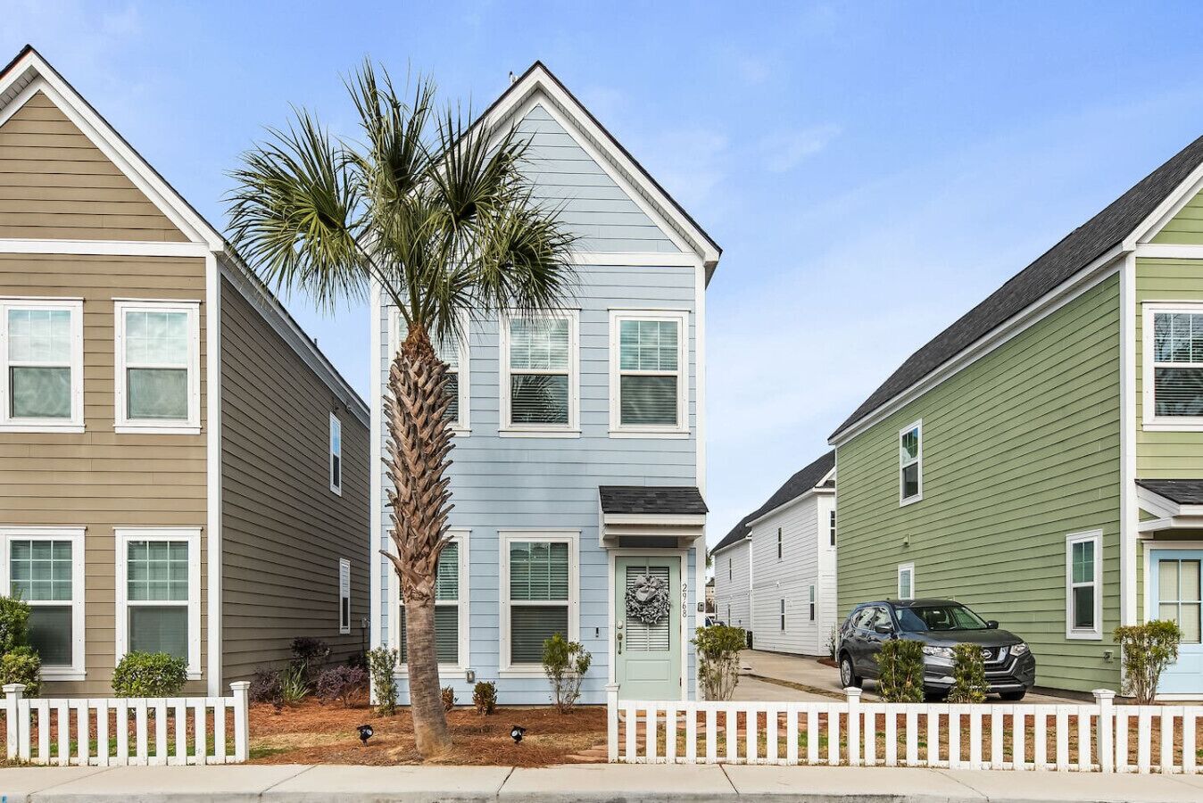 A row of townhouses with a white picket fence, a palm tree, and a car parked in front.