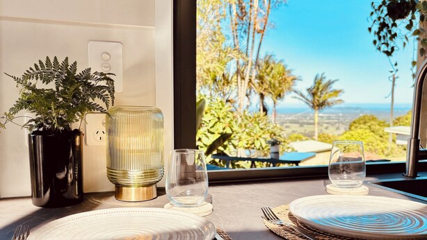 Un comedor con vista a palmeras y un cielo despejado. Hay dos platos, un vaso y una planta en maceta sobre la encimera.