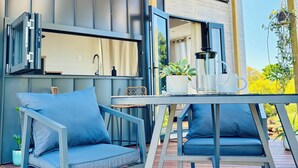 Un patio exterior con piso de madera, dos sillas azules con cojines, una mesa y una planta colgante.