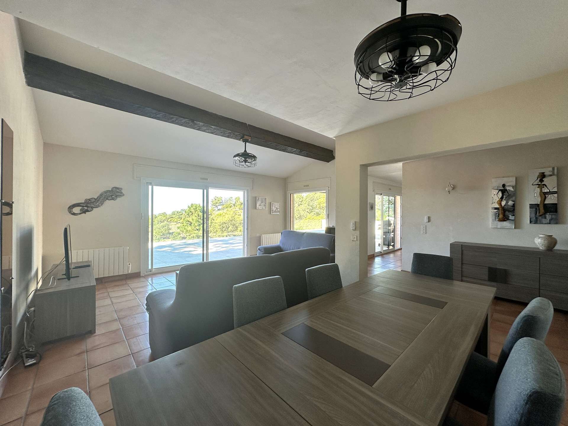 Un vaste espace dédié à la salle à manger, avec une longue table en bois et des chaises modernes, un ventilateur de plafond et une vue sur l’extérieur grâce à de grandes portes coulissantes.