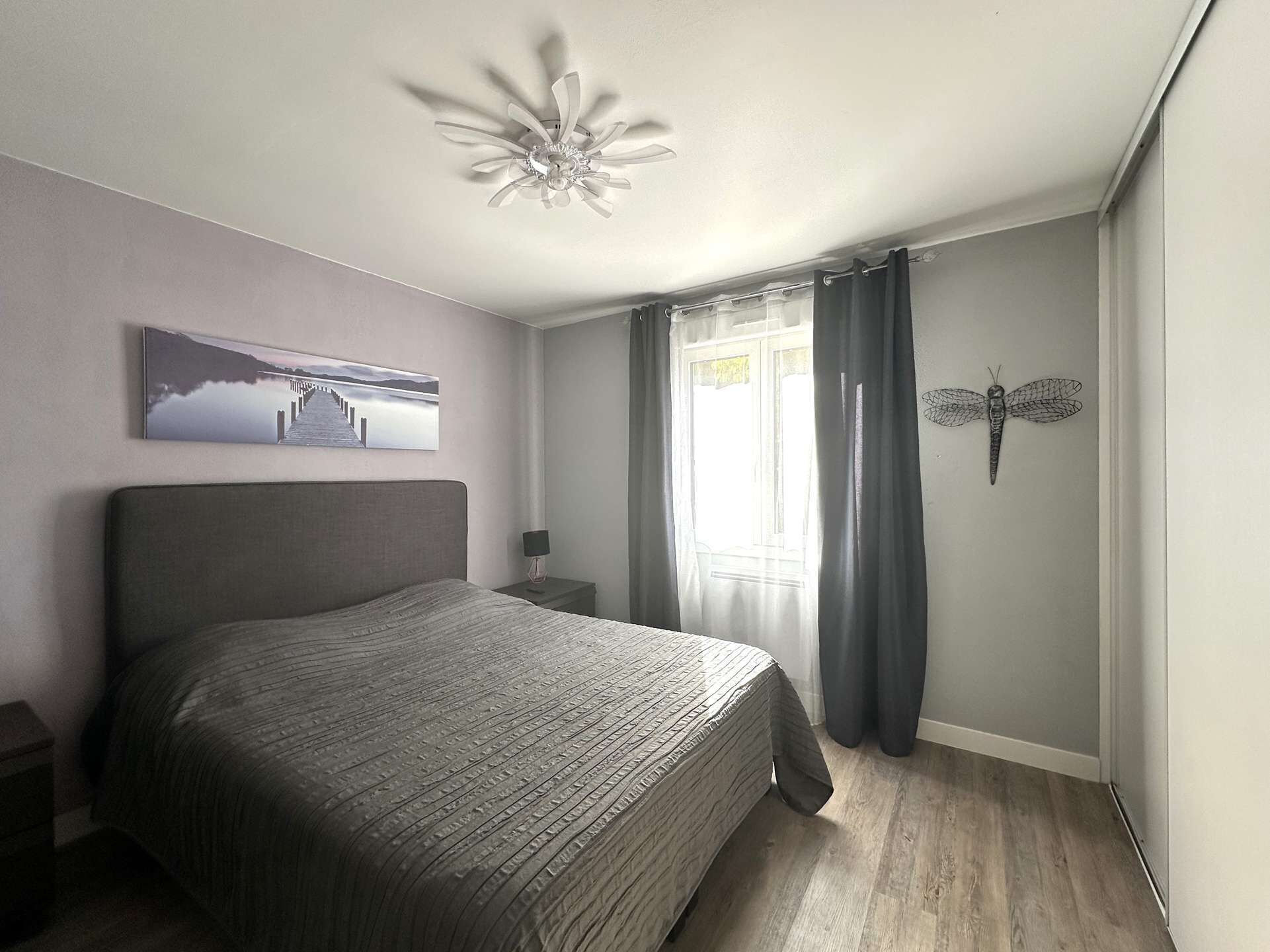Un dormitorio con cama, mesita de noche, una obra de arte en la pared, ventilador de techo y ventana con cortinas.