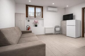 A modern kitchen with a beige sofa, a white refrigerator, a TV on top of the fridge, and a window with lace curtains. - Vespucci Studio - Next to the Sea (Alassio)