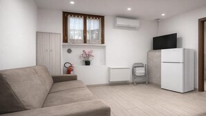 Una cucina moderna con un divano color beige, un frigorifero bianco, una TV sopra il frigo e una finestra con tende in pizzo.
