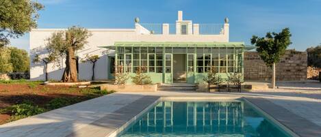 A pool with a glass enclosure, a white building with a green roof, and a stone wall.