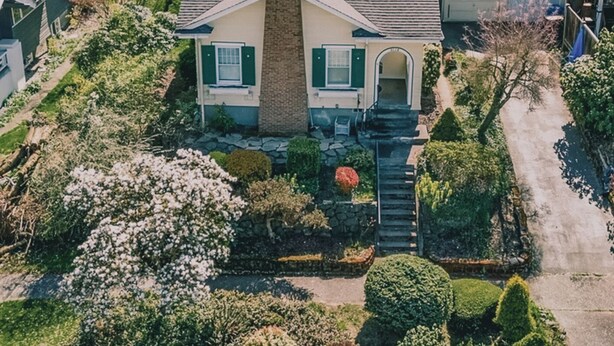 Une maison de plain-pied dotée d’une cheminée imposante, d’un porche à l’avant et d’un jardin bien entretenu où poussent diverses plantes et fleurs.