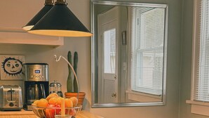 Une cuisine avec un plan de travail en bois, une machine à café, un grille-pain et un saladier rempli de fruits. Il y a une fenêtre avec des stores et un miroir sur le mur.