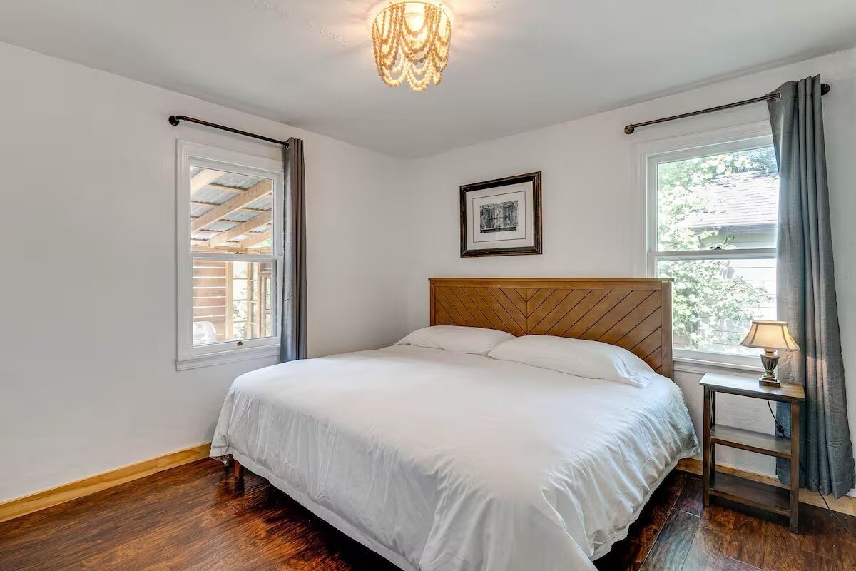 Une chambre à coucher avec une tête de lit en bois, un lit recouvert de linge de lit blanc, une petite table de chevet avec une lampe, un tableau encadré au mur et une fenêtre avec des rideaux.