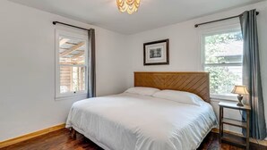 A bedroom with a wooden headboard, a bed with white bedding, a small bedside table with a lamp, a framed picture on the wall, and a window with curtains. - Cozy 1-bedroom cottage in serene Fiddletown (Fiddletown)