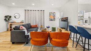 Ett modernt vardagsrum med en grå soffa, orangefärgade stolar, ett litet bord med dekorativa föremål och en utsikt över naturen genom ett fönster.