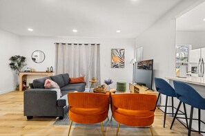A modern living room with a grey sofa, orange chairs, a small table with decorative items, and a view of the outdoors through a window. - Game On, Lights Low - Arcade & Pool Table Nights (Midway City)
