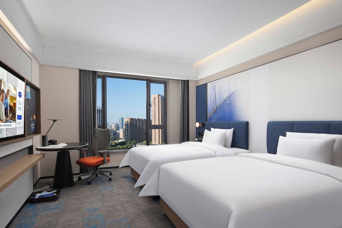 Hotelkamer met twee bedden, een bureau, een stoel en een groot raam met uitzicht over de stad.