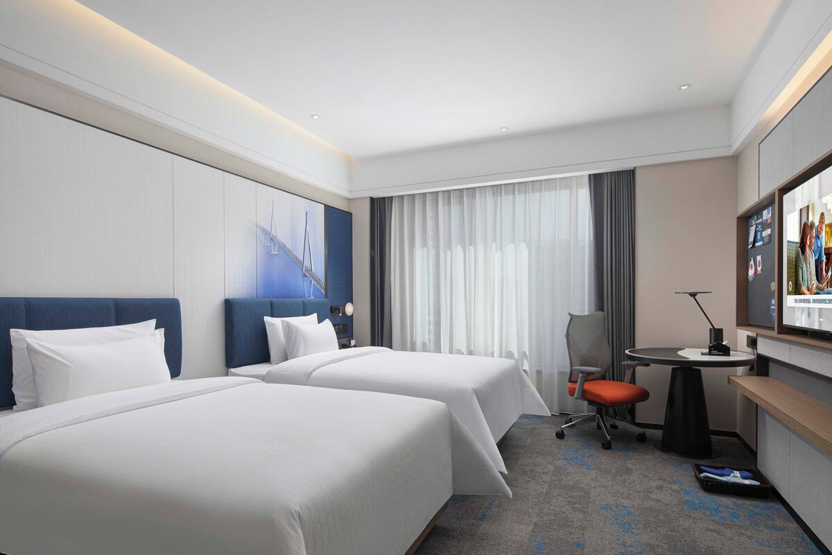 Een hotelkamer met twee bedden, een bureau met stoel, een televisie en een groot raam met gordijnen.