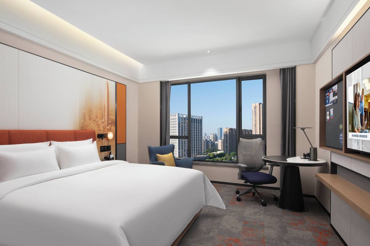 Een hotelkamer met een groot bed, een bureau, een stoel en een televisiescherm waarop een groep mensen te zien is.