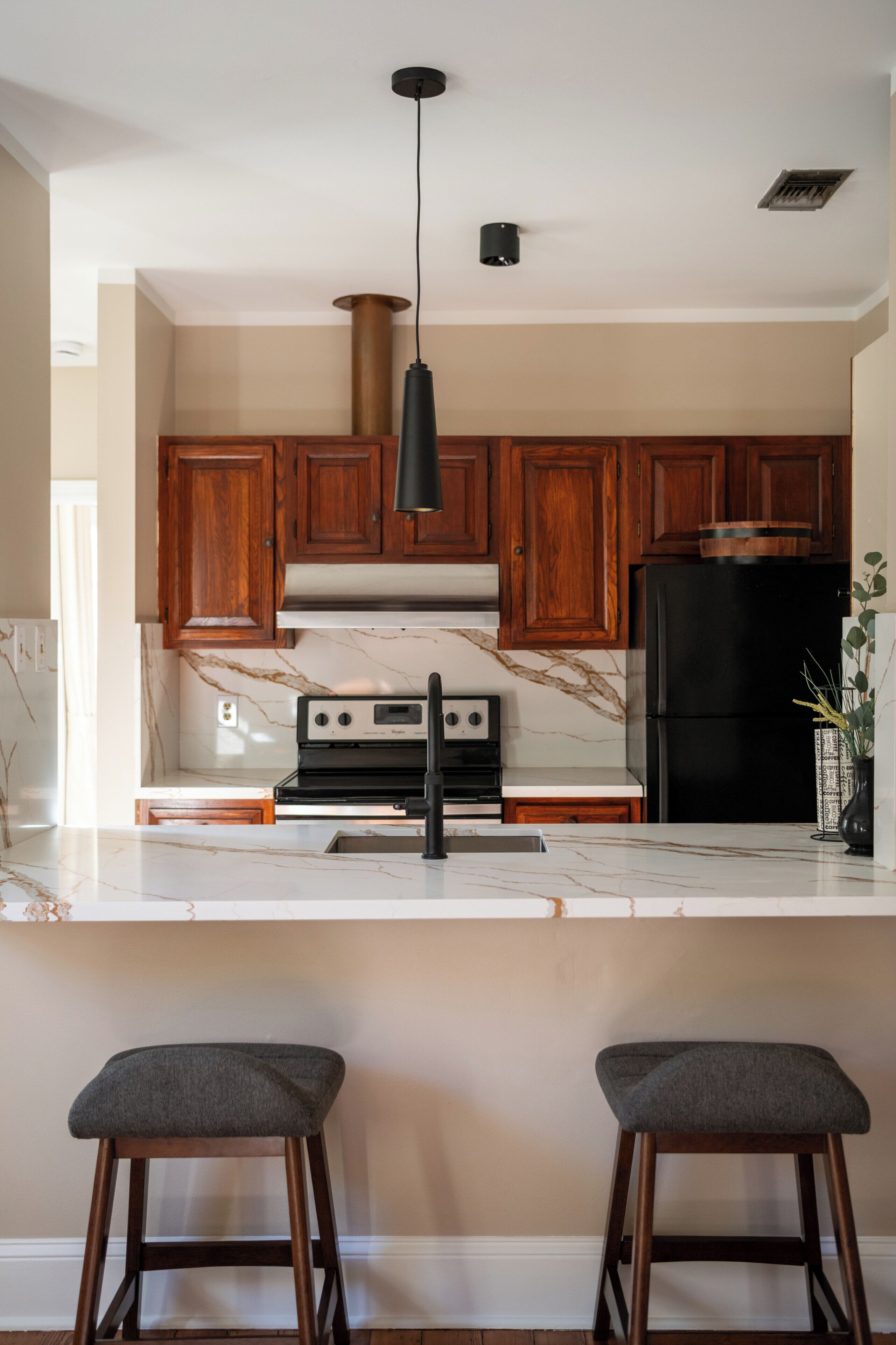 Une cuisine avec des meubles de rangement en bois, un réfrigérateur noir, une cuisinière et deux tabourets de bar.