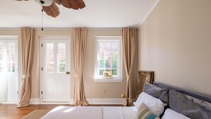 Una camera da letto con un letto grande, tende color beige, una finestra con vista su un edificio in mattoni e una ventola a soffitto.