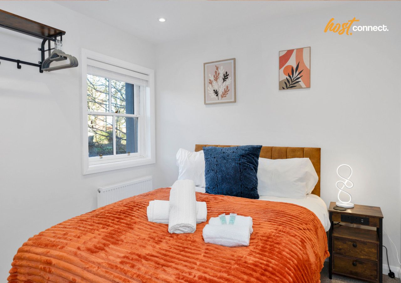 This stylish double bedroom features a plush bed with a vibrant orange throw, modern bedside lighting, and a practical wall-mounted hanging rack. Brightened by natural light, and the room includes fresh white towels.