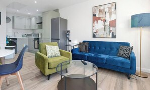 Un salon moderne avec un canapé, un fauteuil et une table basse, une cuisine en arrière-plan et un tableau accroché au mur.