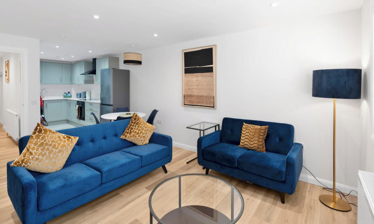 Een moderne woonkamer met twee blauwe banken, een glazen salontafel en een keuken op de achtergrond.