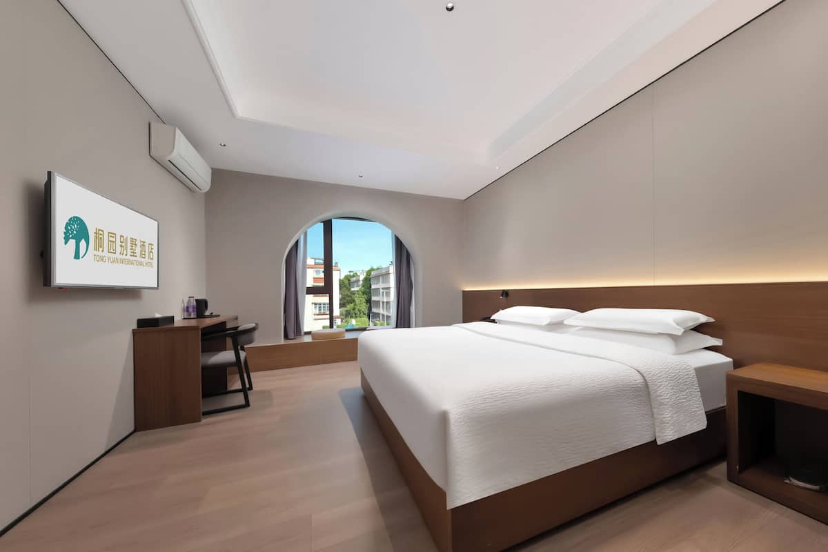 A modern hotel room with a large bed, a desk, and a view through an arched window.