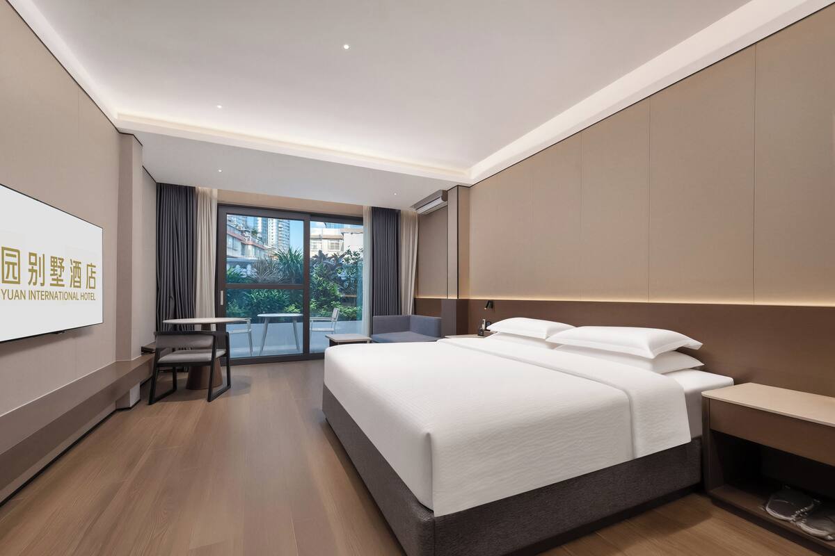 A hotel room with a large bed, a desk, and a chair, a television on the wall, and a view of buildings outside.