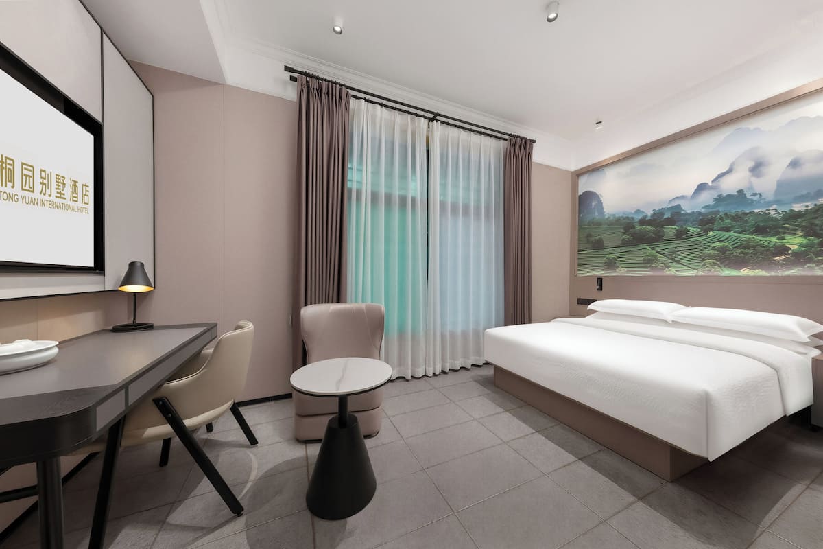 A hotel room with a bed, a desk, a chair, a small table, and a large window with curtains.