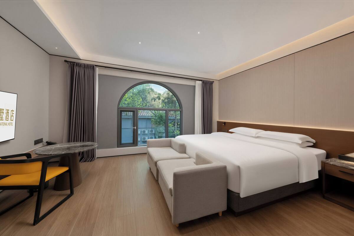 A modern hotel room with a large bed, a desk, a chair, and a view of greenery through a window.
