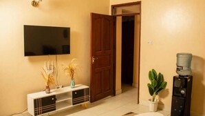 A living room with a flat-screen TV, a wooden door, a water dispenser, and a potted plant. - Paradise Apartments Embu (Embu)